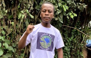 Der Sozial- und Klimaaktivist Daniel Kalalizi steht mit geballter Faust vor Pflanzen mit einem T-Shirt der congo Basin Alliance.