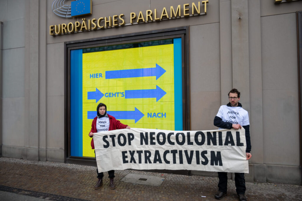 Zwei Personen halten ein Transparent mit der Aufschrift "Stop Neocolonial Extractivism" vor einem Gebäude mit der Aufschrift "Europäisches Parlament".
