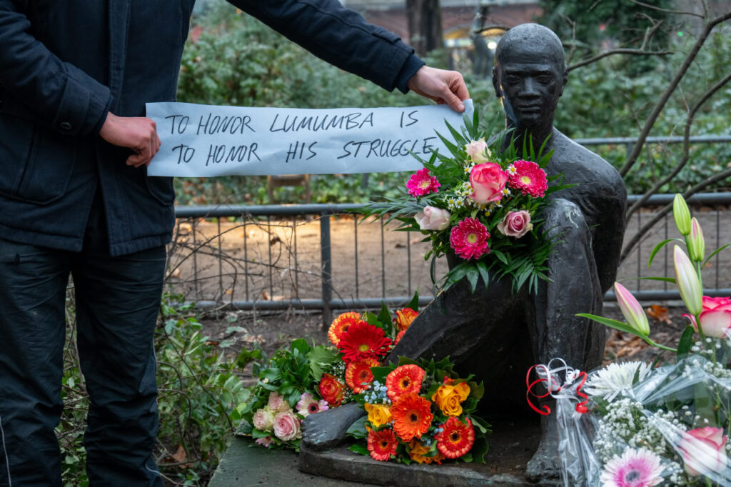 Statue von Patrice Éméry Lumumba von Blumen umgeben. Eine Person hält ein Papier mit einem Spruch neben seinem Kopf.