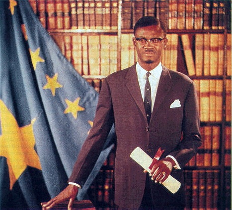 Offizielles Portrait von Patrice Lumumba aus der Zeit als Ministerpräsident. Stehend, die Hand auf einem Tisch stützend (Public Domain), via Wikimedia Commons.